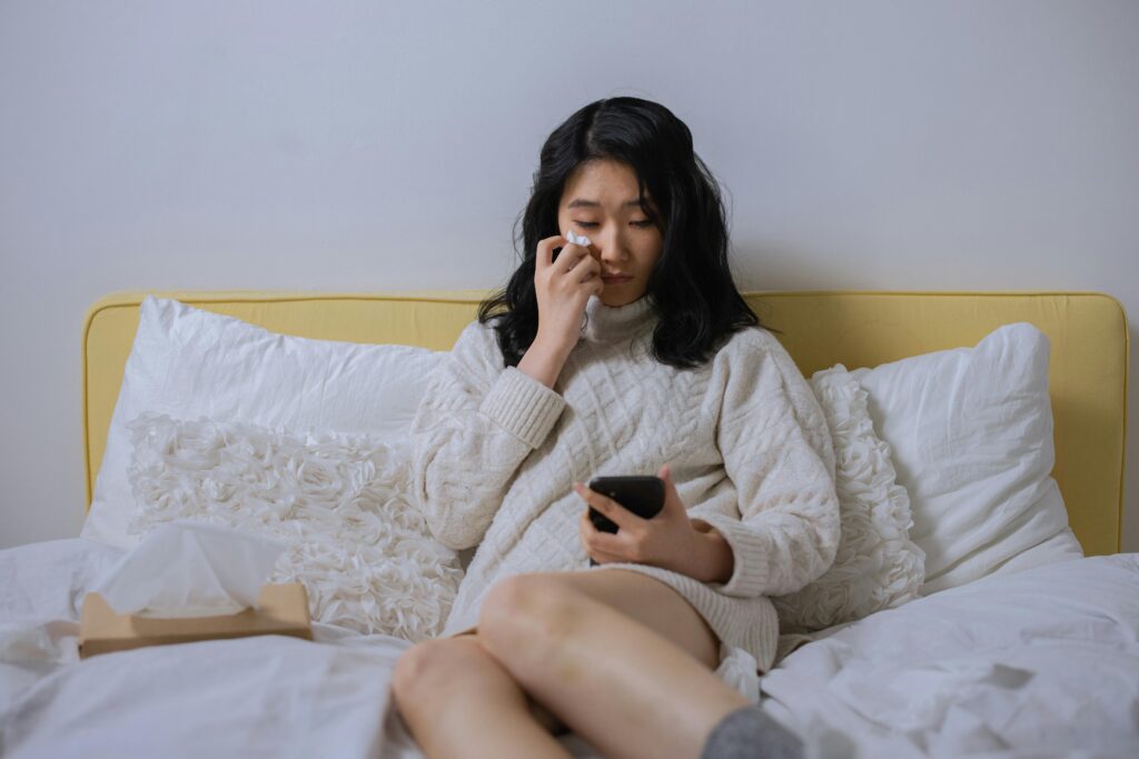 A woman sits on a bed with white sheets, wiping her nose with a tissue while looking at a smartphone in her hand.