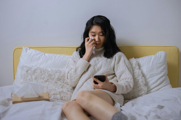 A woman sits on a bed with white sheets, wiping her nose with a tissue while looking at a smartphone in her hand.